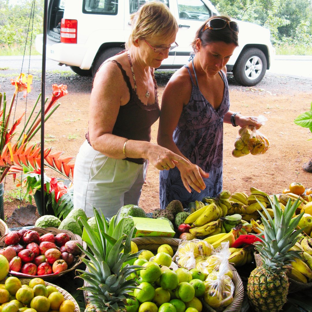 Two women browsing a colourful market fruit stall piled with pineapples, guavas, bananas, apples and tropical flowers