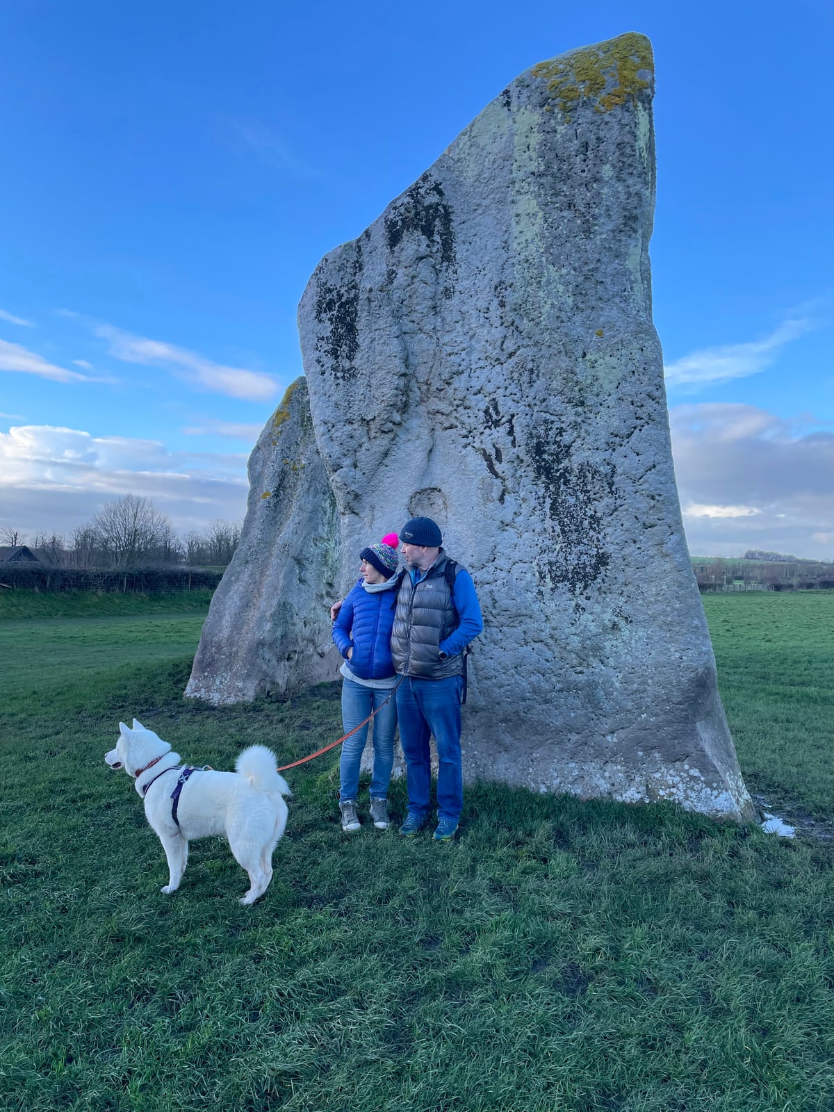 Lisa and Ian with their white dog beside ancient standing stones under a blue sky
