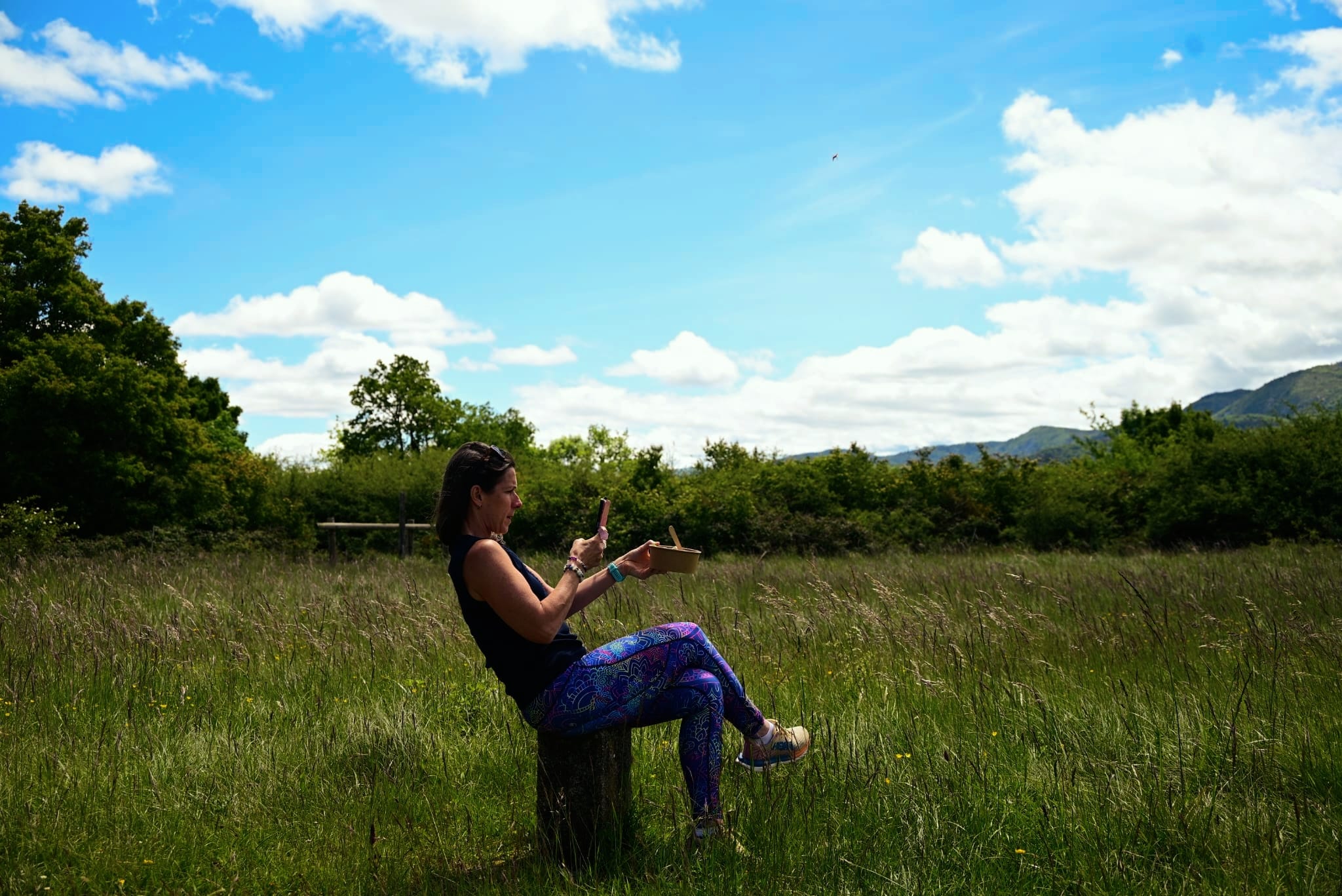 Lisa Picton sitting on a tree stump in a sunlit meadow, using the app over a bowl of food